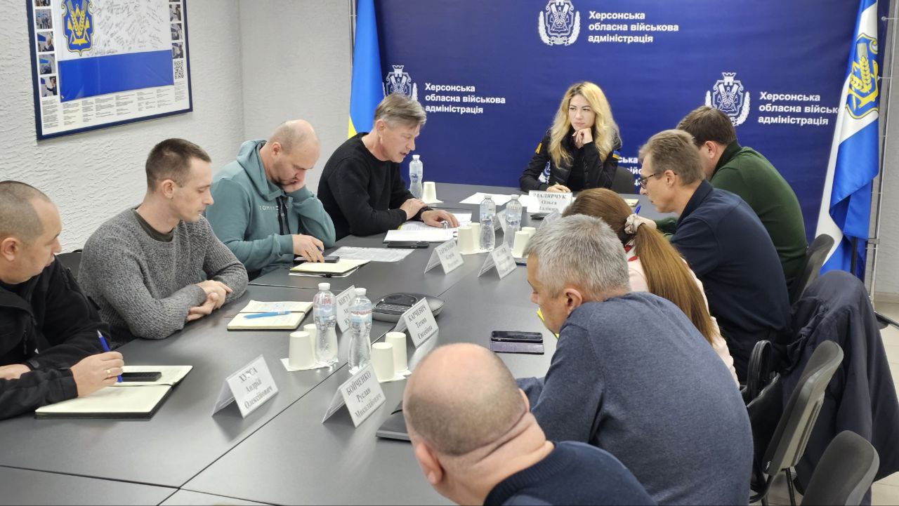 Група людей сидить за довгим столом у приміщенні Херсонської обласної військової адміністрації, беручи участь у нараді.