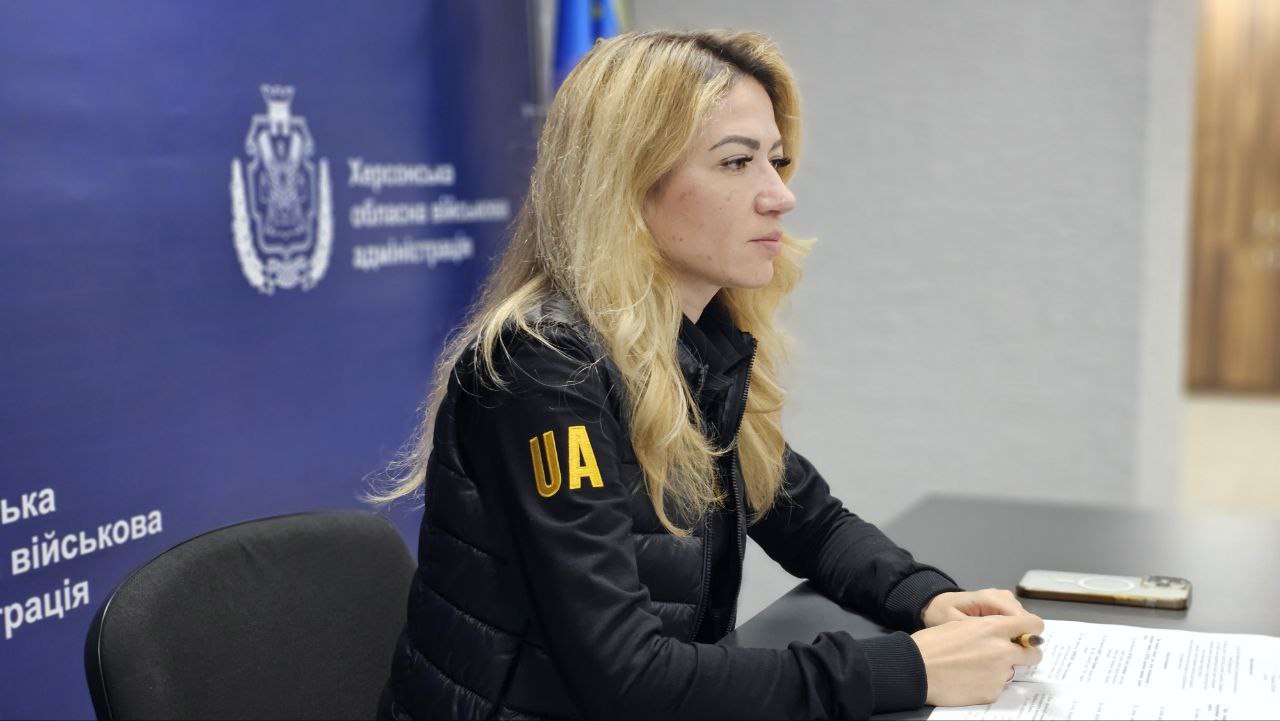 Жінка зі світлим волоссям у чорній куртці з написом: UA сидить за столом, беручи участь у нараді, на тлі символіки Херсонської обласної військової адміністрації.