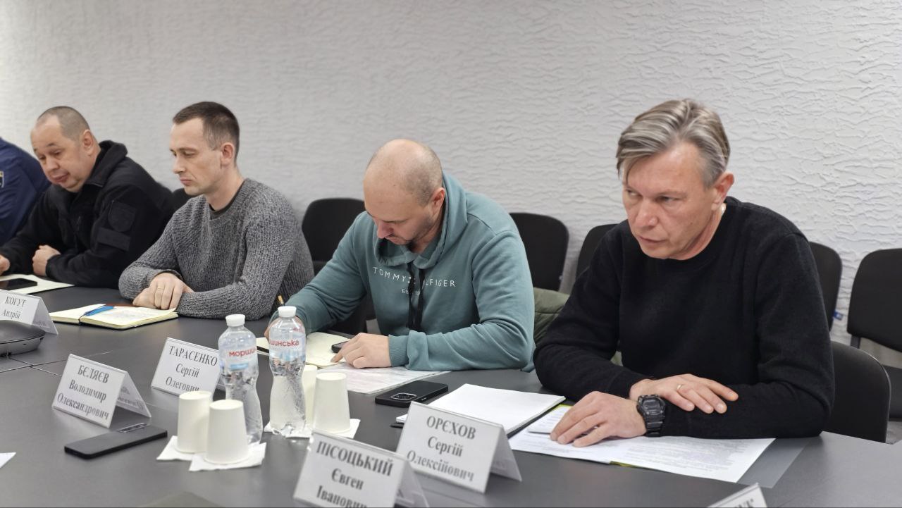  Чотири чоловіки сидять за столом, зосередившись на документах, під час наради. На столі видно їхні іменні таблички.