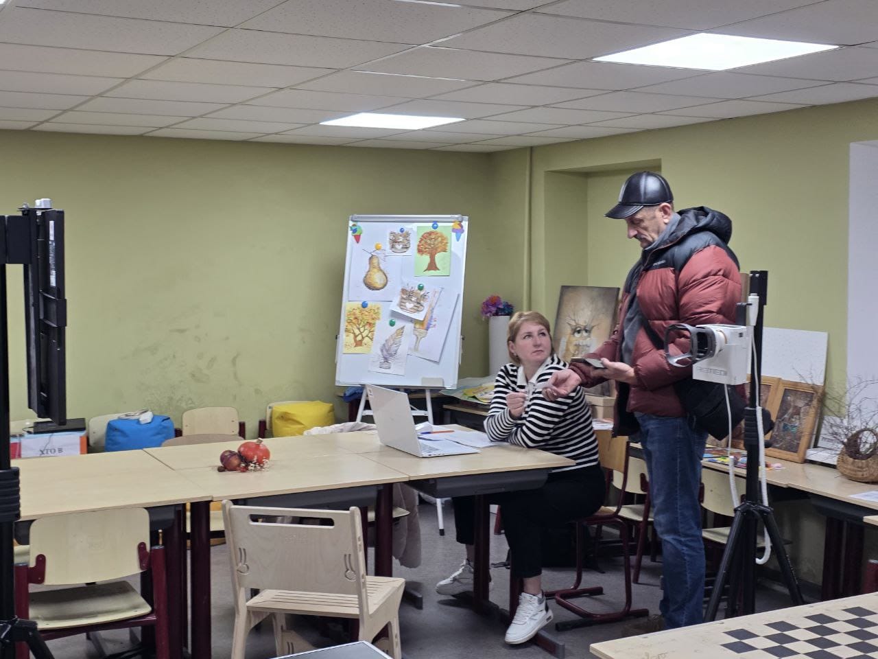 Чоловік у куртці та кепці використовує невеликий пристрій біля руки жінки, яка сидить за столом з ноутбуком у приміщенні, схожому на клас.