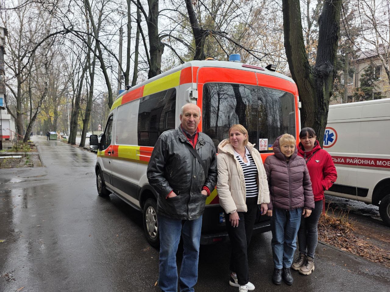 Четверо людей (чоловік і три жінки) стоять на вулиці, позуючи біля задньої частини автомобіля швидкої допомоги або рятувальної служби.