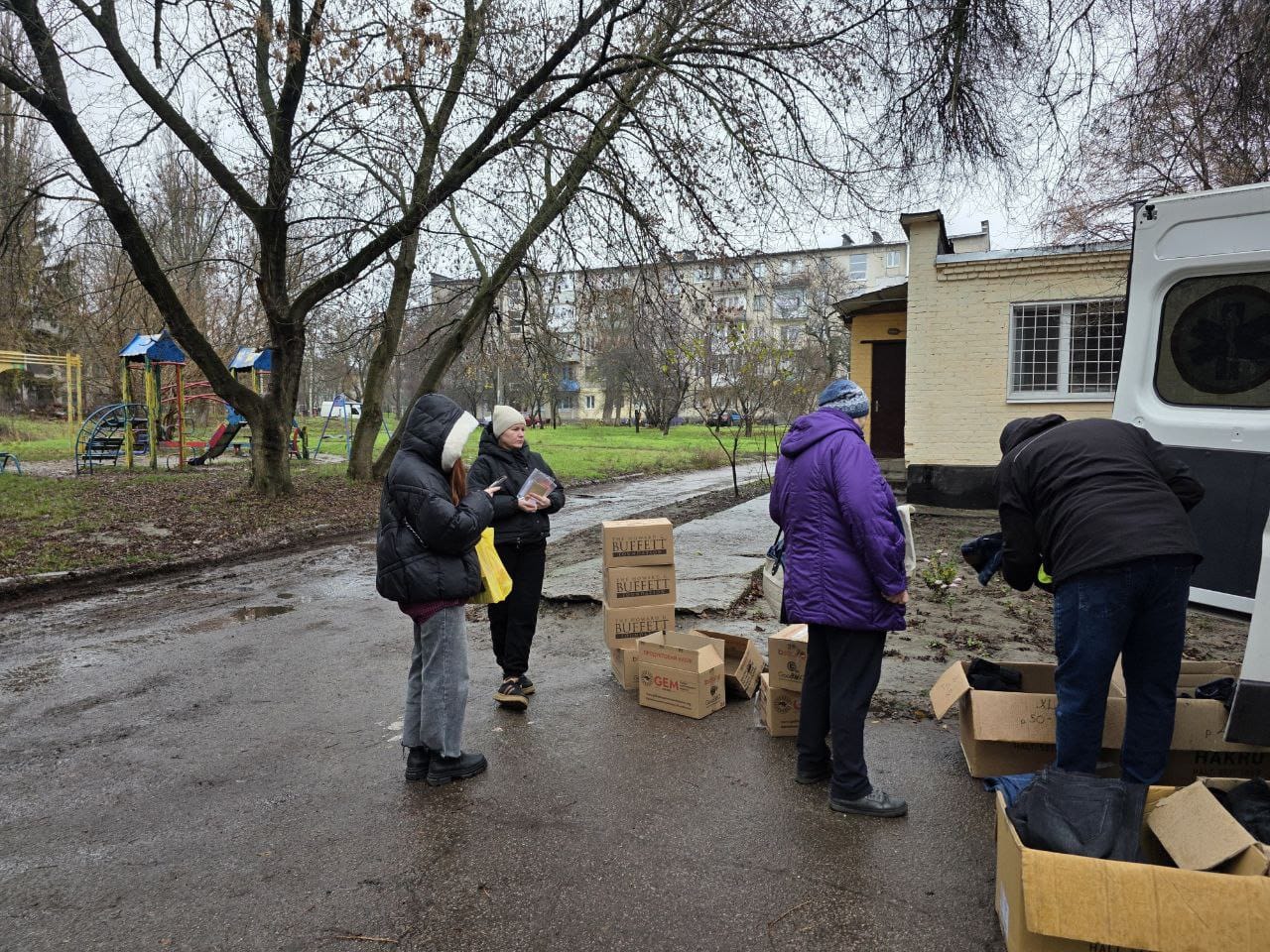  Кілька людей стоять біля стосів картонних коробок, займаючись розвантаженням або роздачею гуманітарної допомоги на вулиці, на тлі житлових будинків.