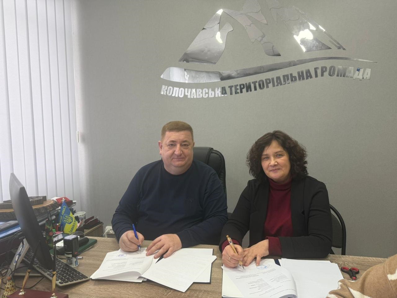 На знімку двоє людей – чоловік у синьому светрі та жінка у бордовій водолазці та чорному піджаку – сидять за офісним столом і одночасно підписують папери. На стіні позаду них видно великий сріблястий логотип та напис: КОЛОЧАВСЬКА ТЕРИТОРІАЛЬНА ГРОМАДА.