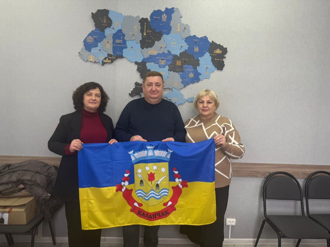 На знімку зображено чоловіка і двох жінок, які стоять поруч і тримають розгорнутий прапор. Прапор має синю та жовту смуги з гербом та написом: КАЛАНЧАК у центрі. На стіні позаду них висить декоративна, багатокольорова дерев'яна мапа України.