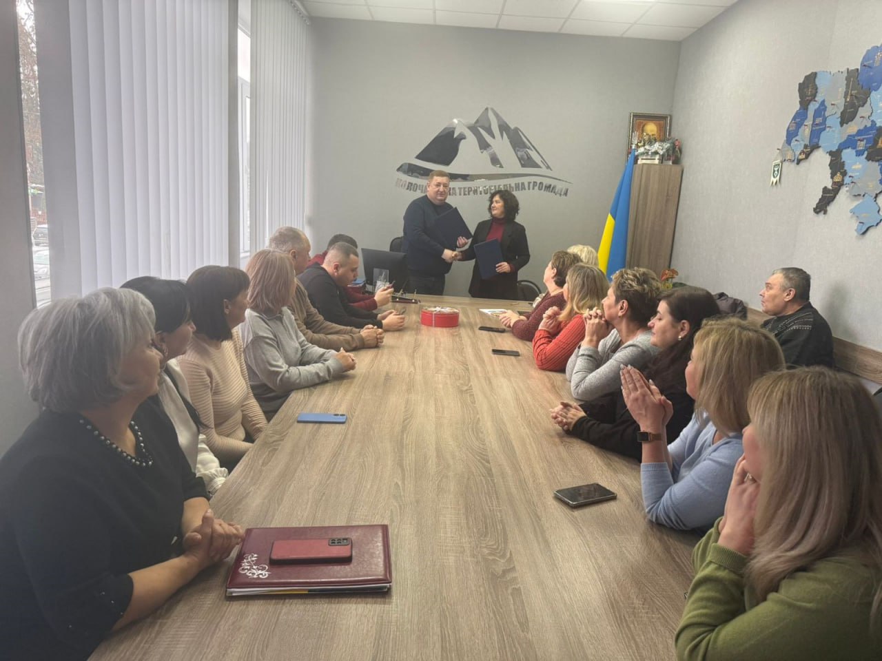 На знімку показано зустріч у приміщенні. Близько п'ятнадцяти людей сидять уздовж довгого дерев'яного столу. У центрі кімнати чоловік і жінка стоять і передають один одному папери. На стіні позаду них видно логотип КОЛОЧАВСЬКА ТЕРИТОРІАЛЬНА ГРОМАДА, прапор України та мапу.