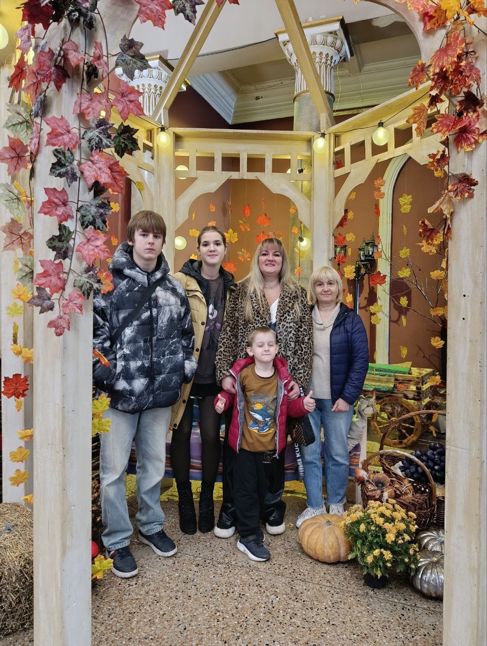 Груповий портрет п'яти людей (три жінки, два хлопці різного віку) у світлій дерев'яній альтанці театру, прикрашеній осіннім листям, гарбузами та кошиками.