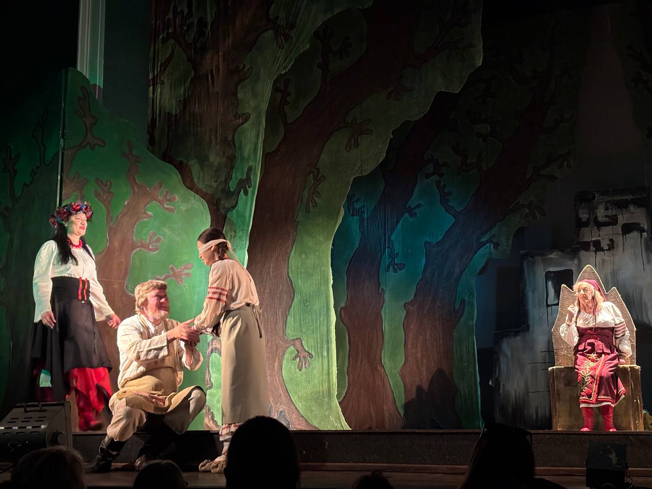 Сцена з театральної вистави: чотири актори в українських народних костюмах на тлі декорацій, що зображують казковий ліс, з великими деревами.