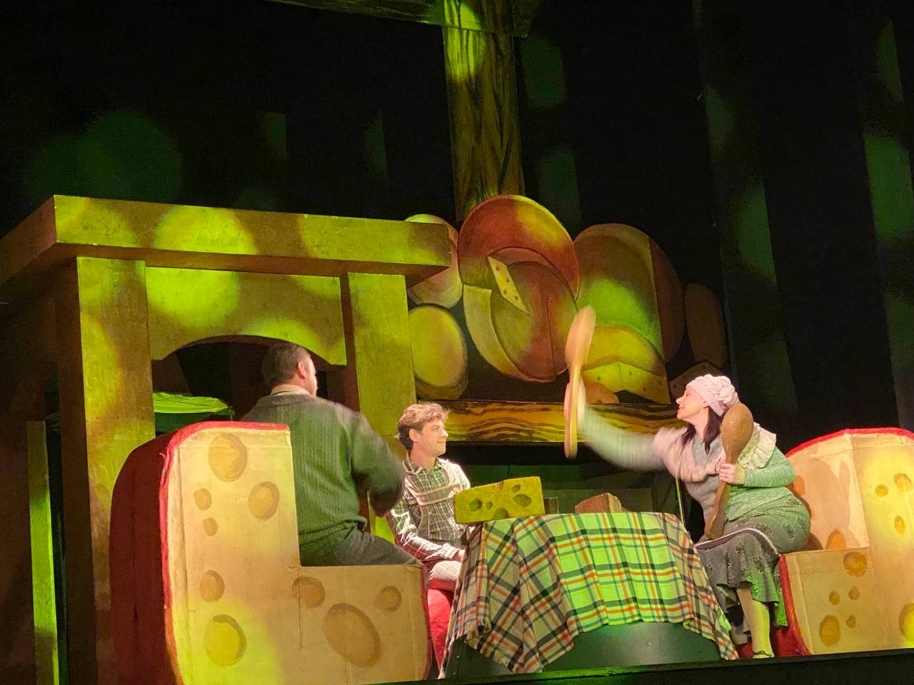 Сцена з дитячої театральної вистави: троє акторів у костюмах сидять за столом, стилізованим під клітинку та сир, з великими шматками сиру як елементами декорацій.