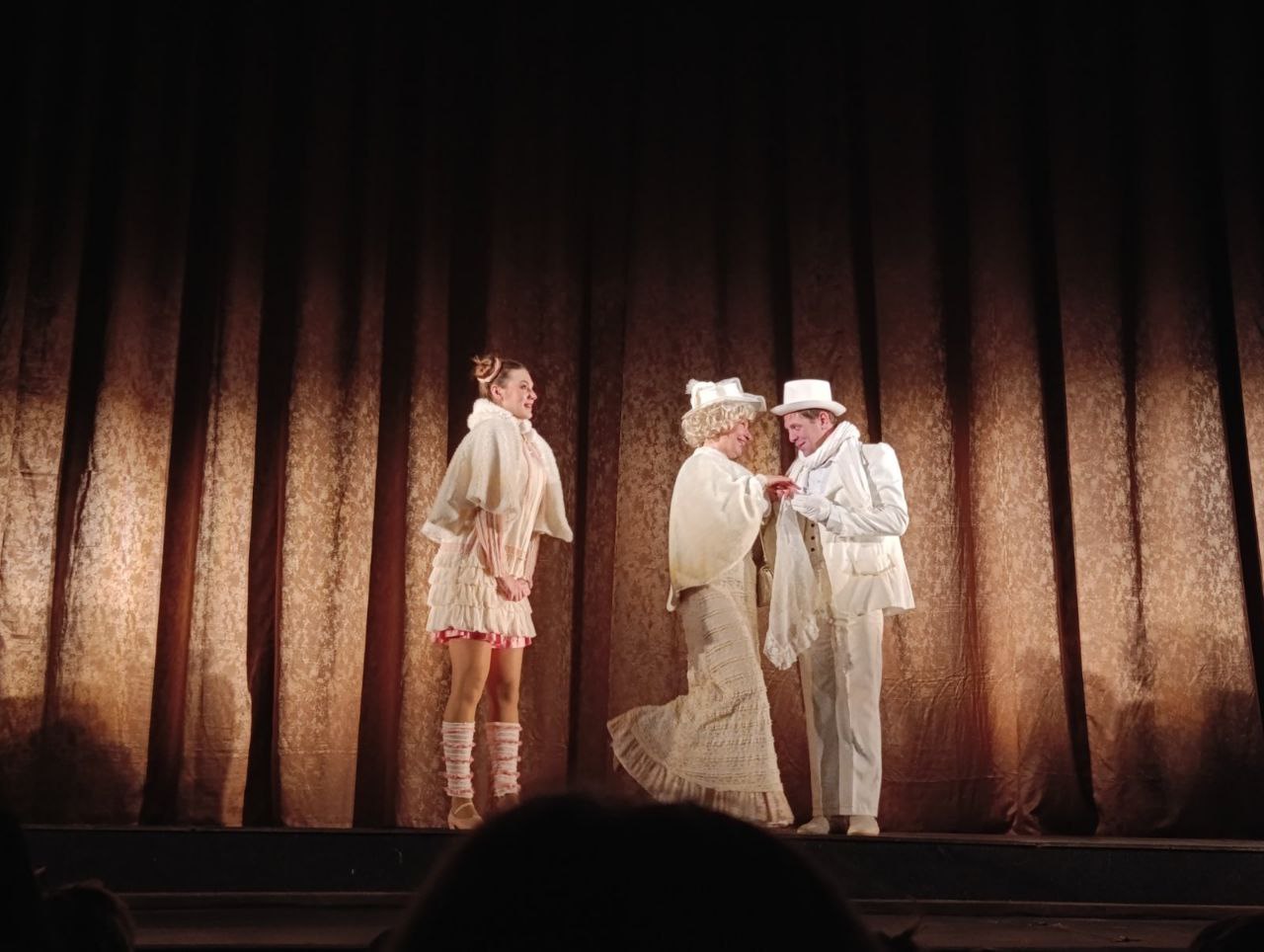 Сцена з театральної вистави: троє акторів у біло-бежевих костюмах на тлі коричневої оксамитової завіси. Пара в центрі в капелюхах взаємодіє, жінка ліворуч стоїть збоку.