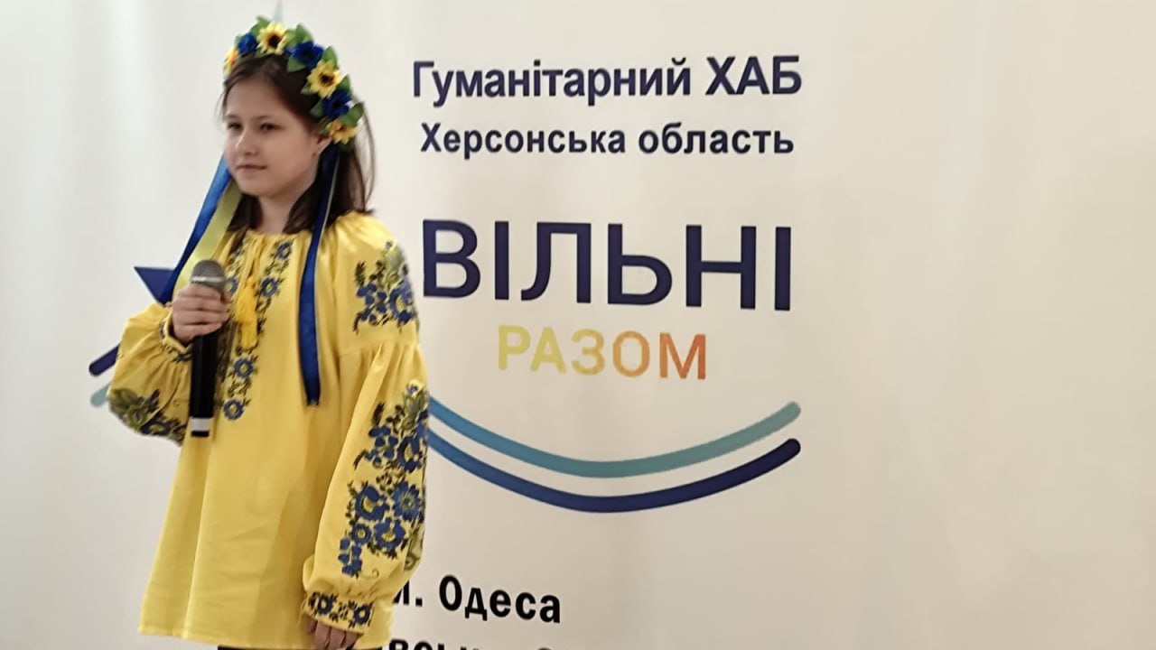 Молода дівчина в яскраво-жовтій вишиванці з синьою вишивкою та українському вінку зі стрічками стоїть перед банером, тримаючи в руці мікрофон. На банері написано: Гуманітарний ХАБ Херсонська область та великими літерами ВІЛЬНІ РАЗОМ.