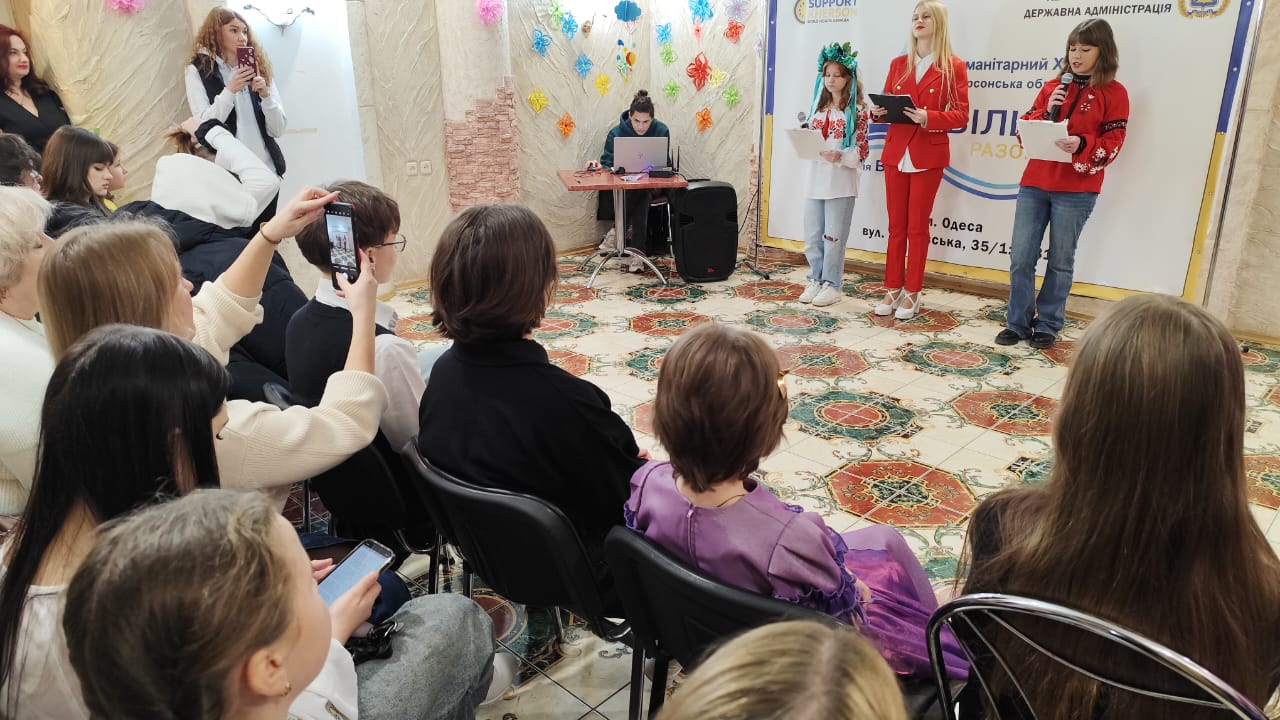 Група людей сидить і слухає виступ на сцені. На сцені чотири молоді жінки, дві з яких тримають папери і виступають, а ще одна в українському вінку. На задньому плані видно банер з написом: Гуманітарний ХАБ та ВІЛЬНІ РАЗОМ. Деякі глядачі фотографують виступ на телефони.