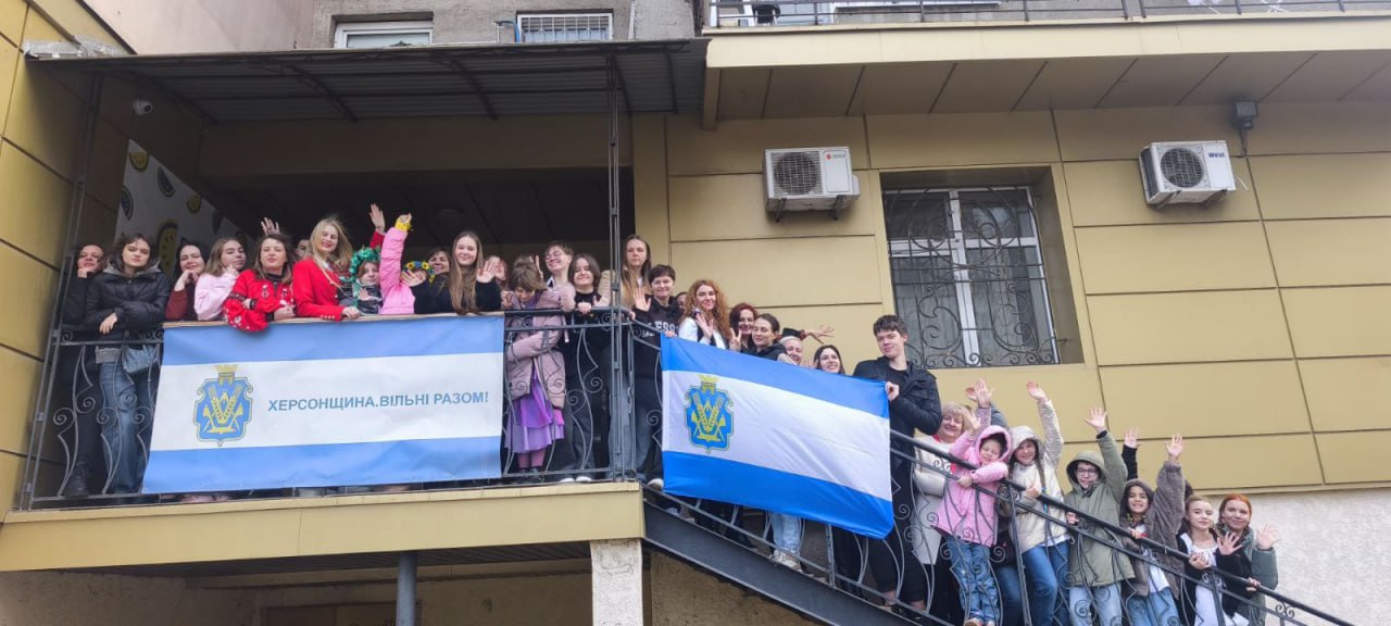 На знімку велика група усміхнених дівчат і кількох хлопців махають руками, стоячи на балконі та зовнішніх сходах двоповерхової будівлі. На балконі висить прапор Херсонської області з написом: ХЕРСОНЩИНА. ВІЛЬНІ РАЗОМ!, а інший такий самий прапор тримають люди на сходах.