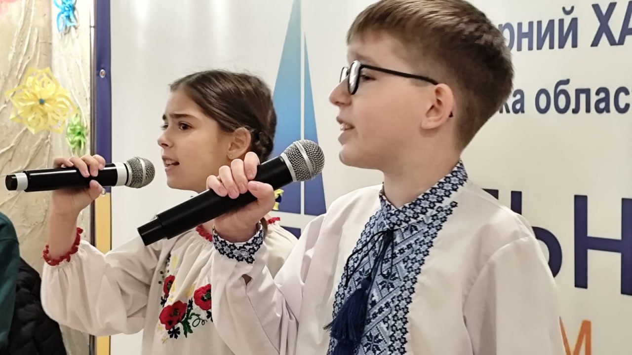 На знімку крупним планом зображено хлопчика та дівчинку, одягнених у традиційні українські вишиванки, які виконують пісню або декламують вірш, тримаючи мікрофони. Хлопчик носить окуляри. На задньому плані видно частину банера з написом українською мовою.