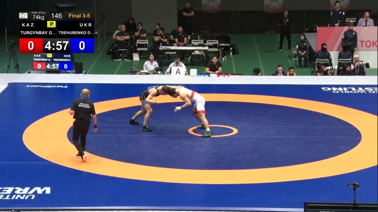 Змагання з вільної боротьби: два спортсмени на килимі, з рефері поруч; на табло відображається рахунок 0-0 і час.