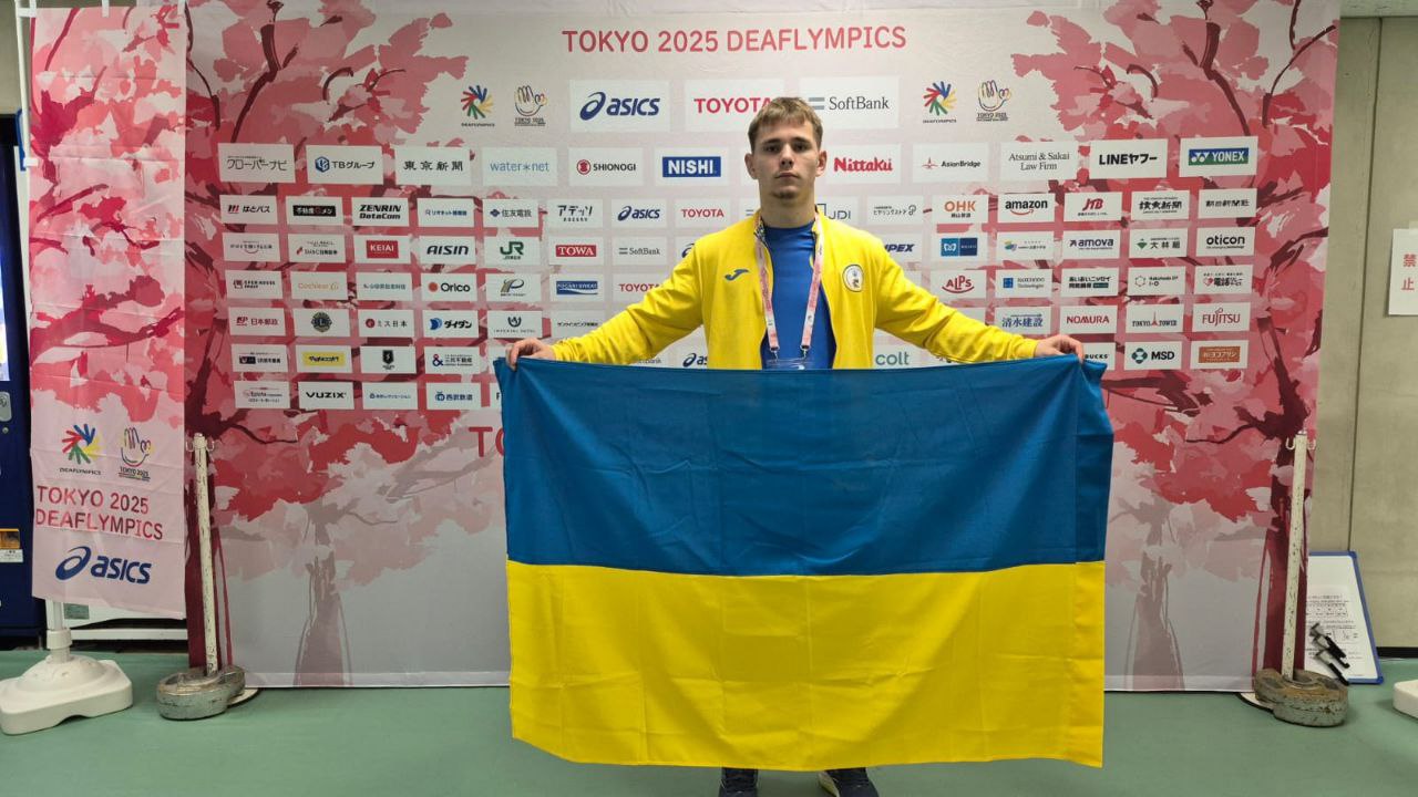 Український спортсмен у спортивній формі стоїть перед спонсорським банером, тримаючи великий прапор України.