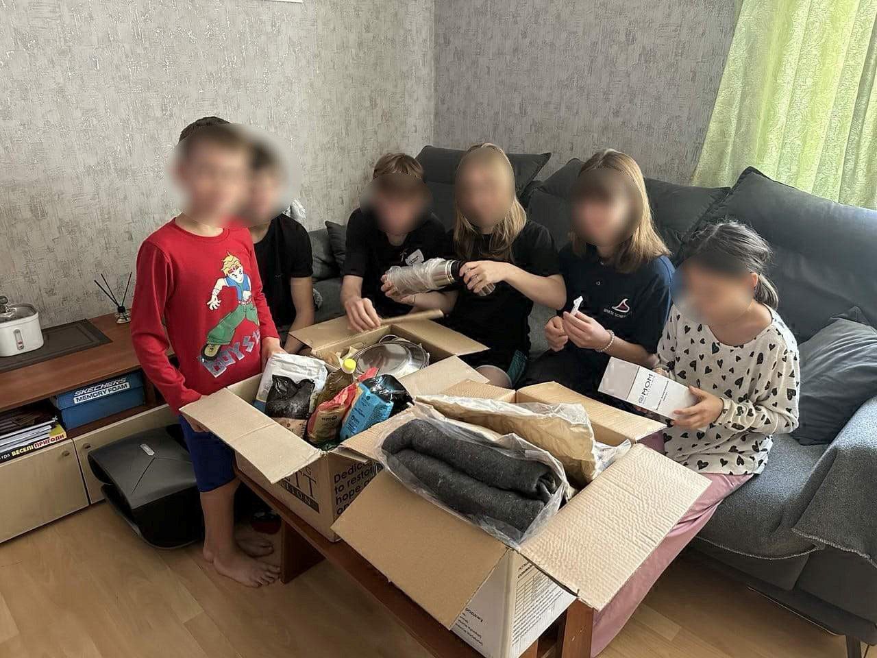 На знімку п'ятеро дітей сидять на дивані, а ще одна дитина стоїть зліва, розбираючи вміст великої картонної коробки, наповненої продуктами, ковдрами та іншими предметами. Внутрішнє оздоблення кімнати включає сірий диван і світло-зелені штори.