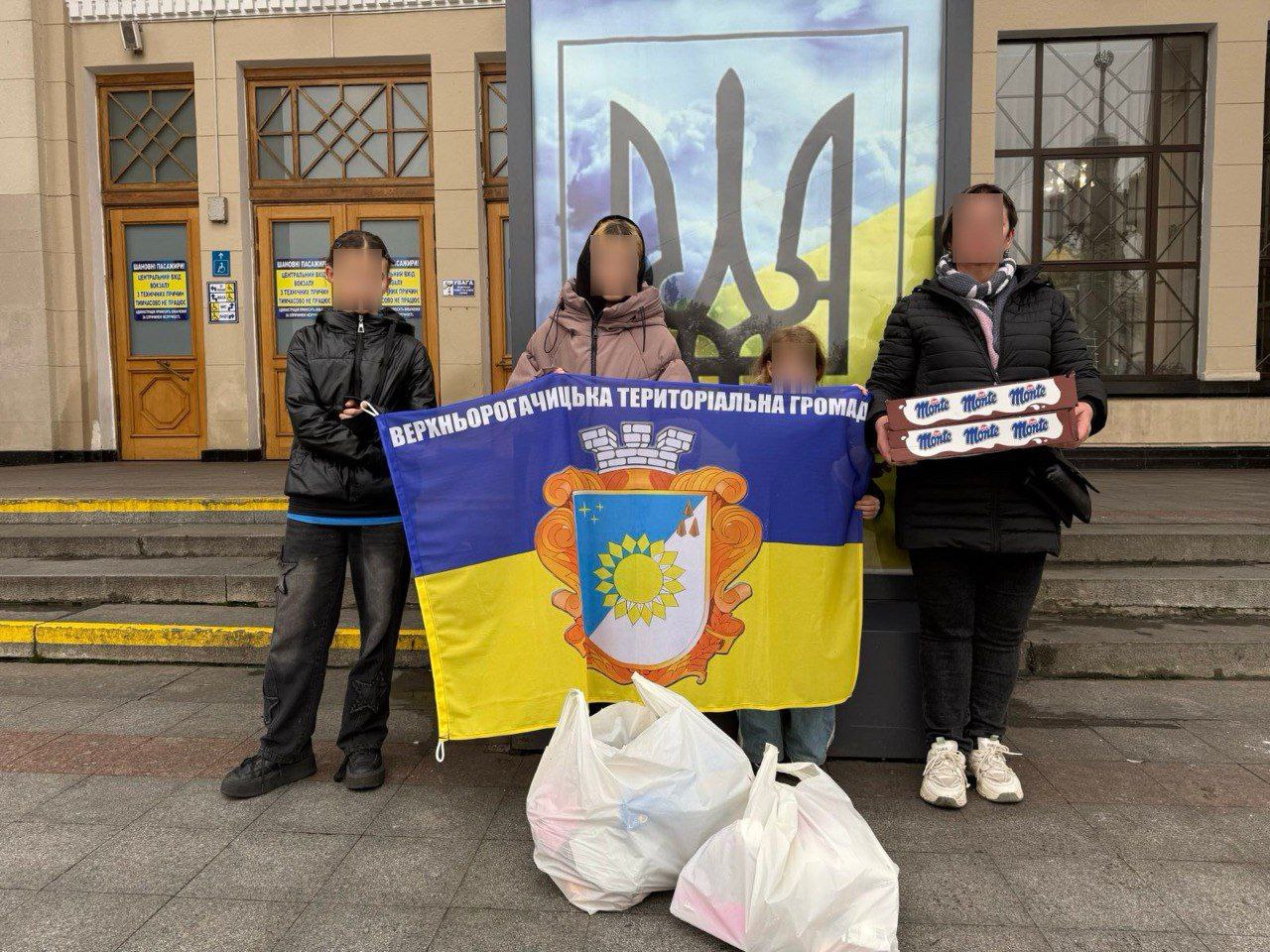 На знімку зображено трьох людей (двох дорослих та дитину), які тримають розгорнутий синьо-жовтий прапор з гербом та написом: ВЕРХНЬОРОГАЧИЦЬКА ТЕРИТОРІАЛЬНА ГРОМАДА. Перед ними на асфальті стоять кілька великих білих пакетів. За ними розташований вхід у будівлю та великий банер із зображенням Тризуба на жовто-синьому тлі.