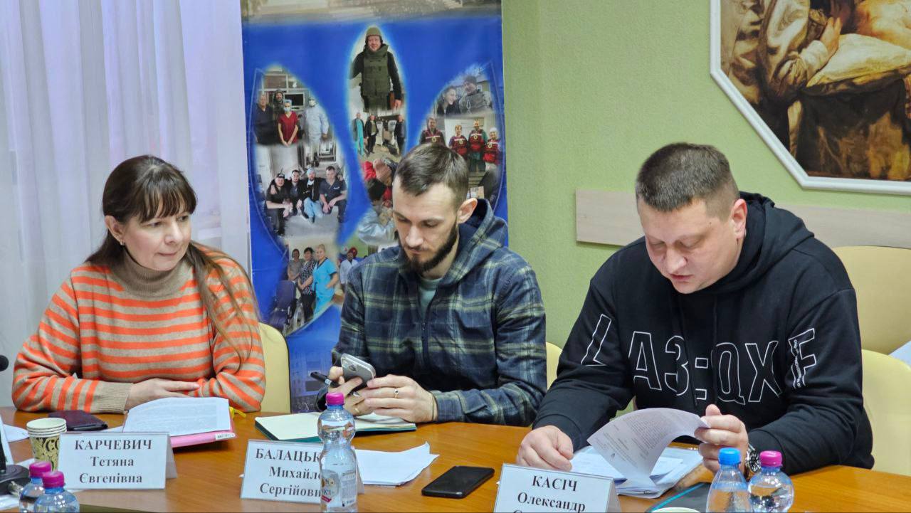 На знімку крупним планом зображено трьох людей за столом: жінка у помаранчево-смугастому светрі, чоловік у картатій сорочці, який дивиться у свій телефон, та чоловік у чорному худі, який читає папери. За ними висить плакат із колажем фотографій людей у військовій та робочій формі.