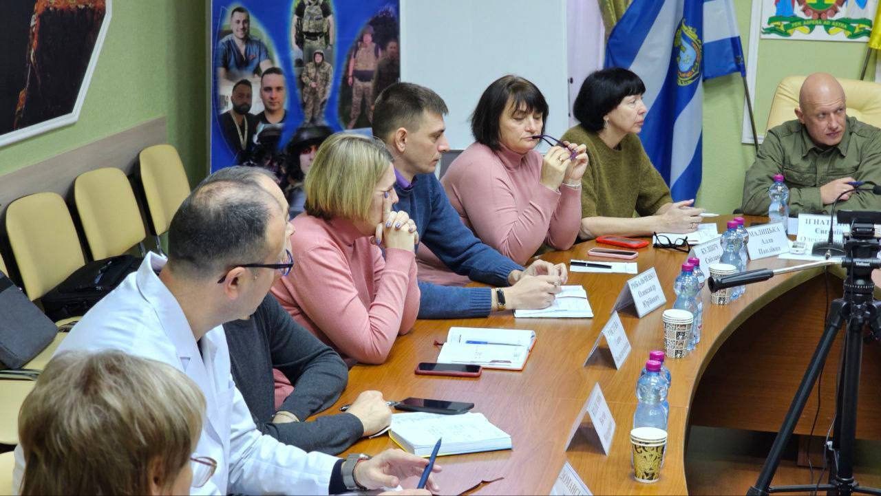 На знімку зображено близько восьми осіб, які беруть участь у діловій зустрічі. Вони сидять уздовж вигнутого дерев'яного столу, на якому лежать блокноти, телефони та пляшки з водою. На дальньому кінці столу праворуч сидить лисий чоловік у зеленій військовій сорочці. На задньому плані видно плакати, прапори та герб.