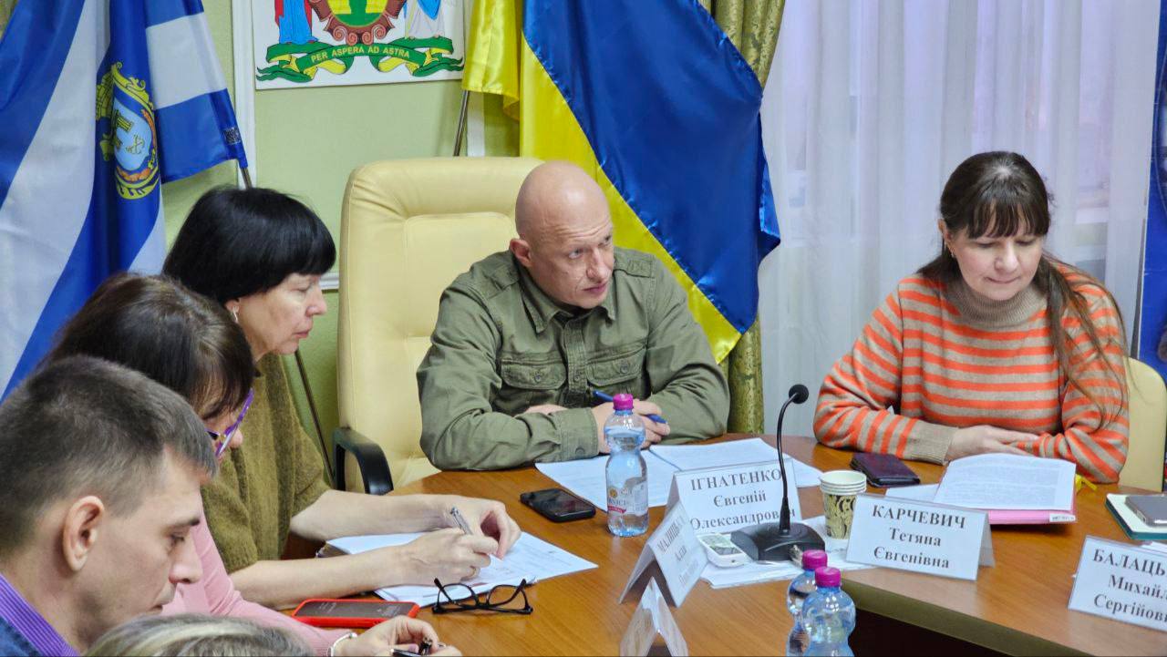 На знімку зображено частину ділової зустрічі. У центрі – лисий чоловік у зеленій сорочці, який говорить, та жінка у смугастому светрі. Перед ними розташовані таблички з іменами. На задньому плані видно прапори (зокрема, прапор України) та герб.