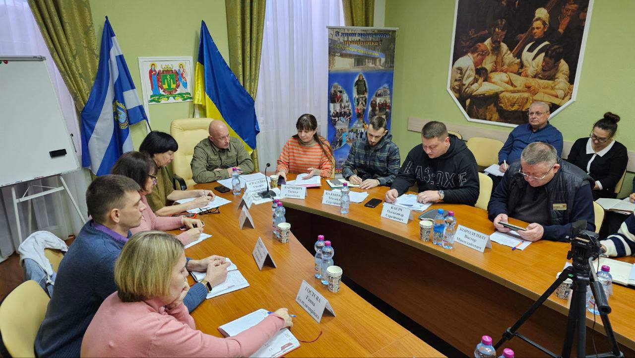 На знімку показано ділову зустріч у приміщенні. Група людей, включаючи чоловіків та жінок, сидить навколо U-подібного дерев'яного столу. На столі видно пляшки з водою та таблички з іменами. На задньому плані розташовані прапори (один український), плакати та картина.