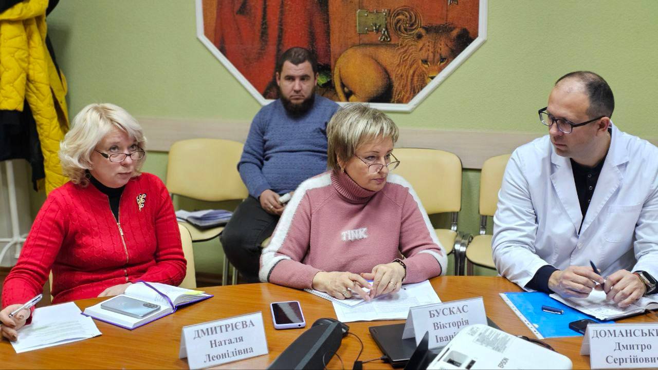 На знімку крупним планом зображено трьох учасників наради, які сидять за дерев'яним столом. Зліва – жінка у червоному светрі, в центрі – жінка у рожевому светрі, а справа – чоловік в окулярах і білому медичному халаті. На задньому плані сидить ще один чоловік.