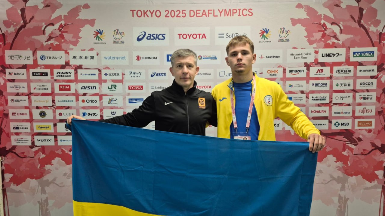 Двоє чоловіків, тренер та спортсмен у жовто-синій формі, тримають великий прапор України на тлі пресволу зі спонсорами та логотипом Tokyo 2025 Deaflympics.