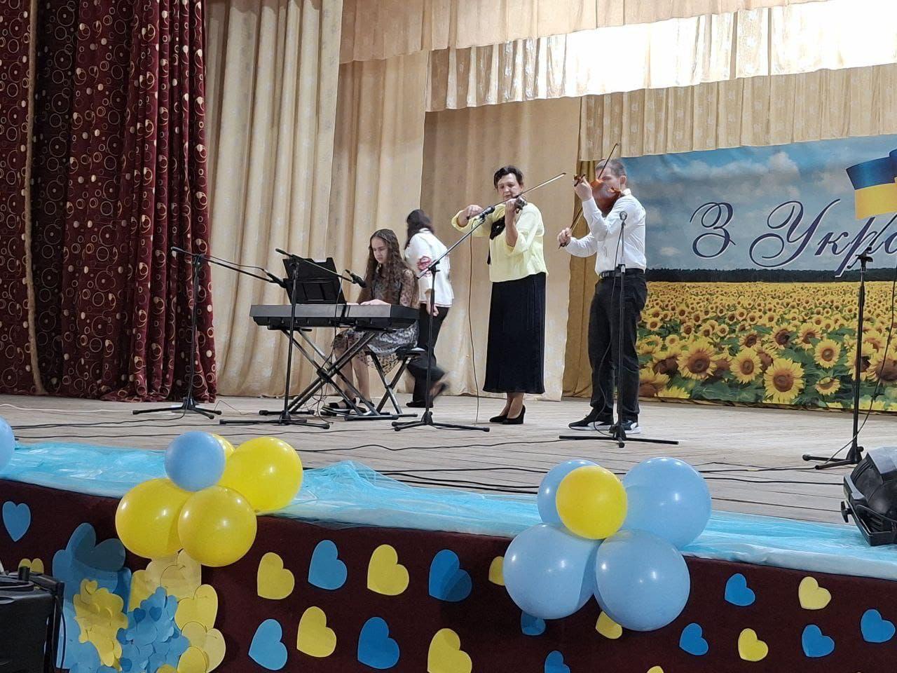 Музичний виступ на сцені. Жінка та чоловік грають на скрипках, а дівчина — на синтезаторі. Сцена прикрашена синьо-жовтими кульками та великим фоном із соняшниками та написом: З Україною в серці!