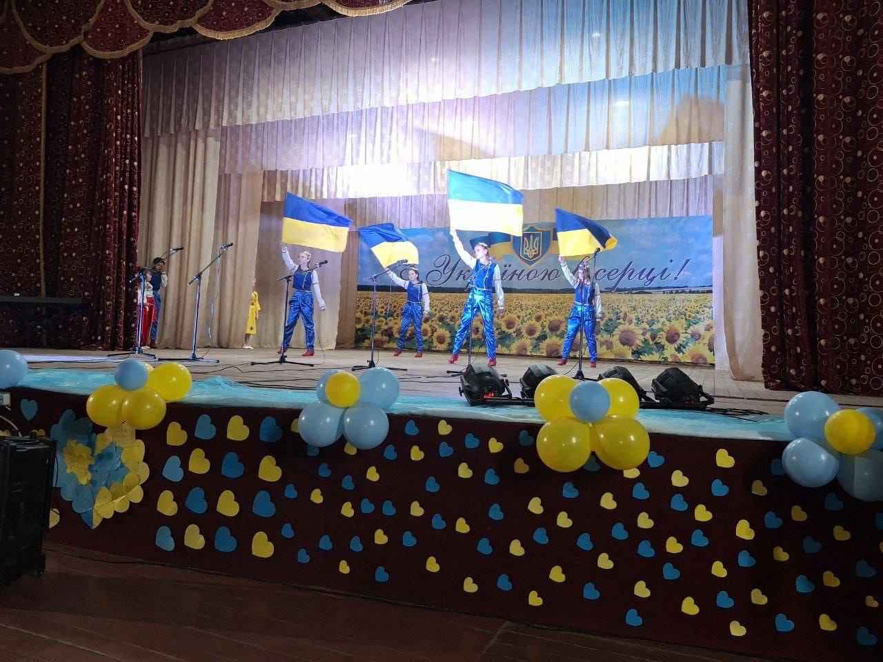 Діти або молодь у синіх сценічних костюмах виступають на сцені, тримаючи українські прапори. Задній фон прикрашений зображенням соняшникового поля та написом: З Україною в серці! Сцена оздоблена синьо-жовтими кульками та сердечками.