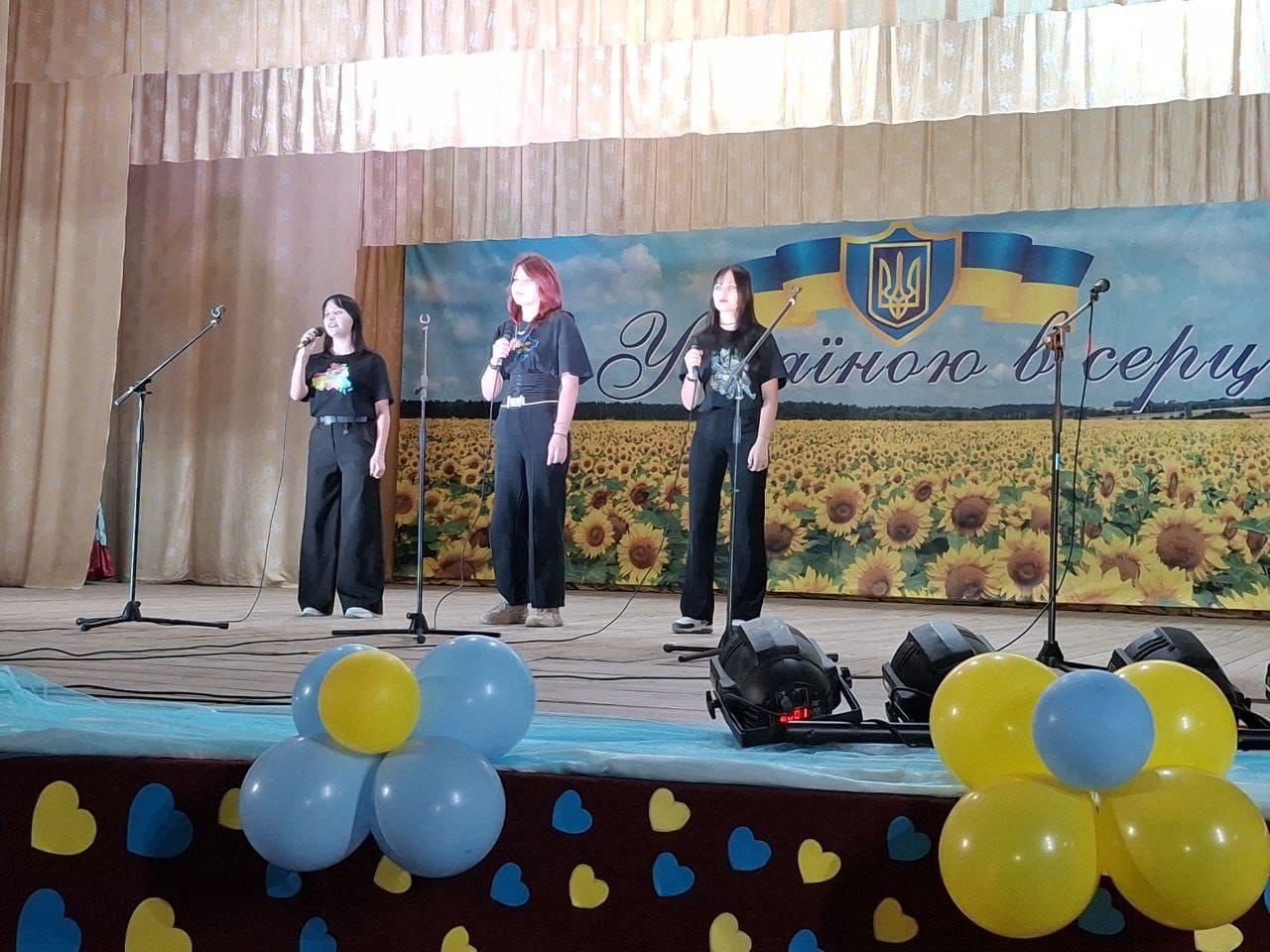 Три дівчини стоять на сцені перед мікрофонами, співаючи або виступаючи. На задньому плані розташований великий банер із соняшниковим полем, українським прапором і написом: З Україною в серці. Сцена прикрашена синіми та жовтими кульками.