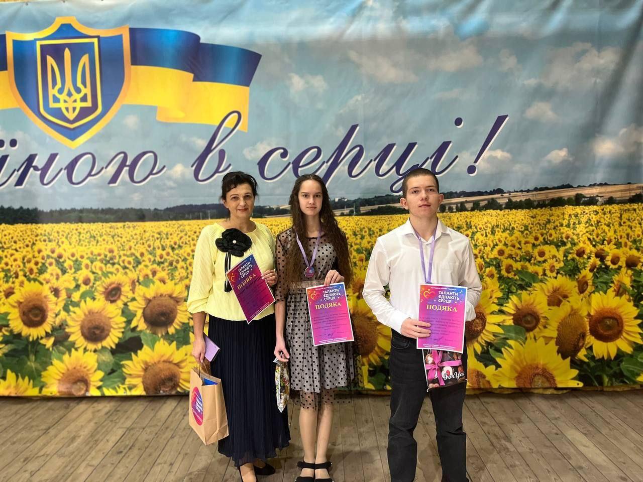 Троє людей — доросла жінка, дівчина та хлопець — стоять разом, тримаючи нагородні подяки, на тлі великого банера з українським прапором, гербом (Тризубом), соняшниковим полем та написом, що закінчується словами: З Україною в серці!