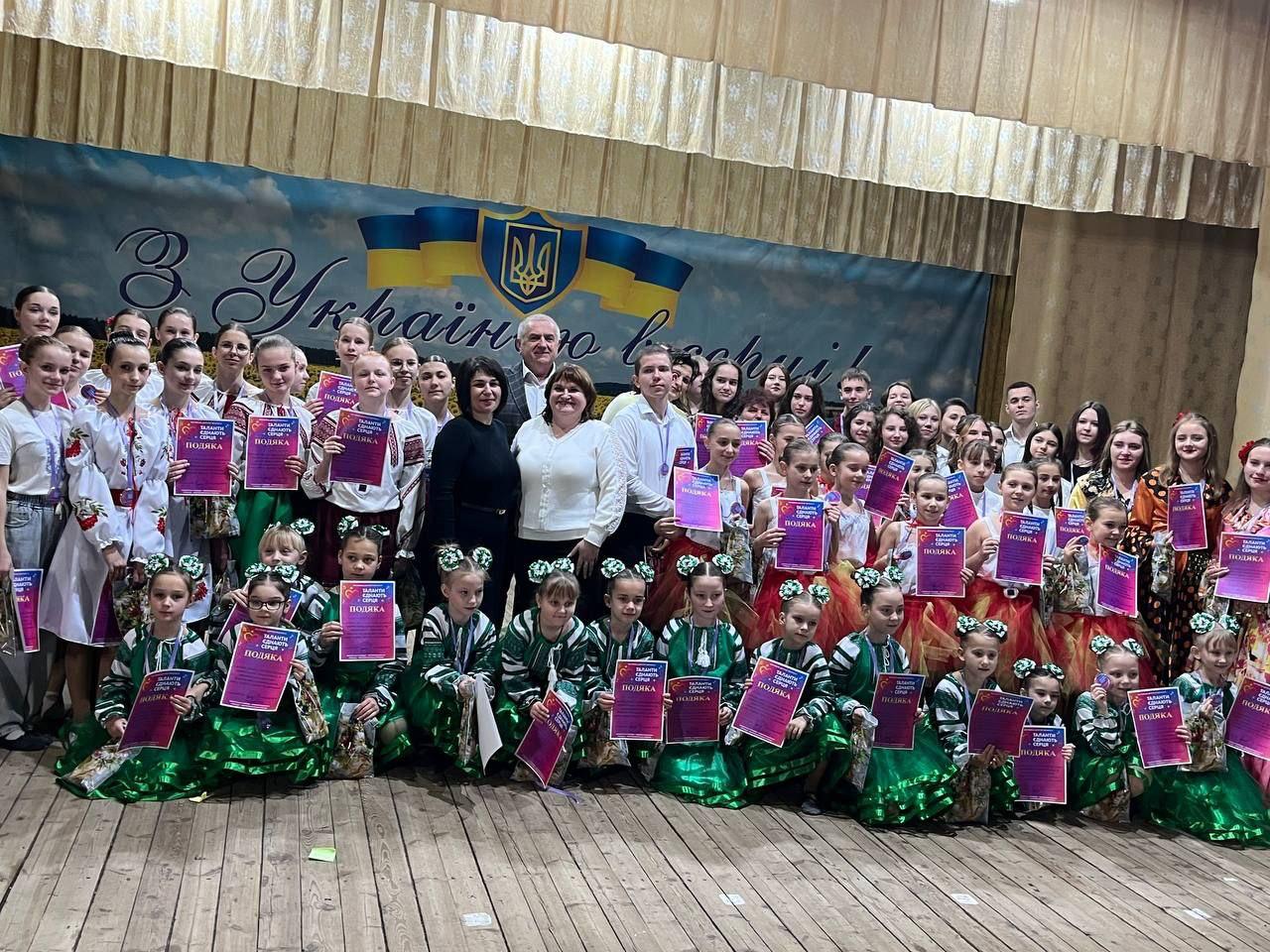 Велика група дітей та підлітків у сценічних та народних костюмах, а також кілька дорослих, позують на сцені, тримаючи нагородні подяки. На задньому плані банер з українським прапором, Тризубом та написом: З Україною в серці!
