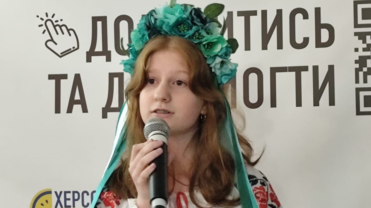 Дівчина з рудим волоссям, у вінку з бірюзовими та зеленими квітами та у вишиванці, виступає з мікрофоном на тлі стіни з частиною напису: Додатись та допомогти.