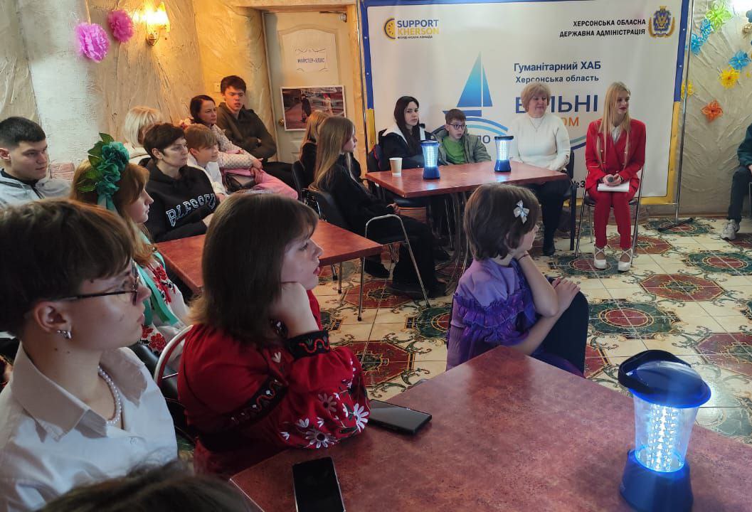 Група молоді та дорослих сидить за столами, слухаючи виступ у приміщенні Гуманітарного хабу Херсонської області Вільні, на задньому плані видно банер з логотипом Херсонської ОДА.