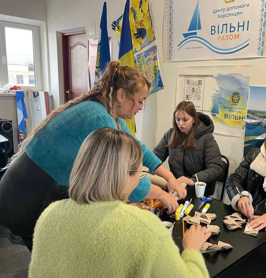 Кілька жінок займаються рукоділлям за столом у волонтерському центрі Вільні разом (Центр допомоги Херсонцям, м. Івано-Франківськ), на задньому плані видно українські прапори та агітаційні матеріали.