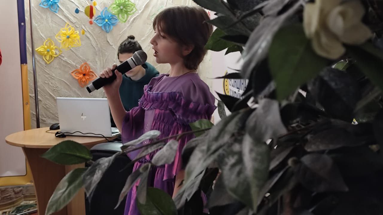Дівчинка з коротким темним волоссям у бузковій сукні співає або виступає з мікрофоном. На задньому плані видно стіну, прикрашену різнокольоровими паперовими квітами, а на передньому плані — гілки великої зеленої рослини.