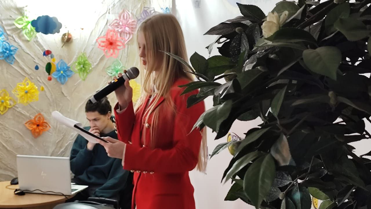 Дівчина з довгим світлим волоссям у яскраво-червоному піджаку виступає з мікрофоном, читаючи з паперу, на тлі стіни, прикрашеної паперовими квітами. Праворуч видно велику зелену кімнатну рослину.