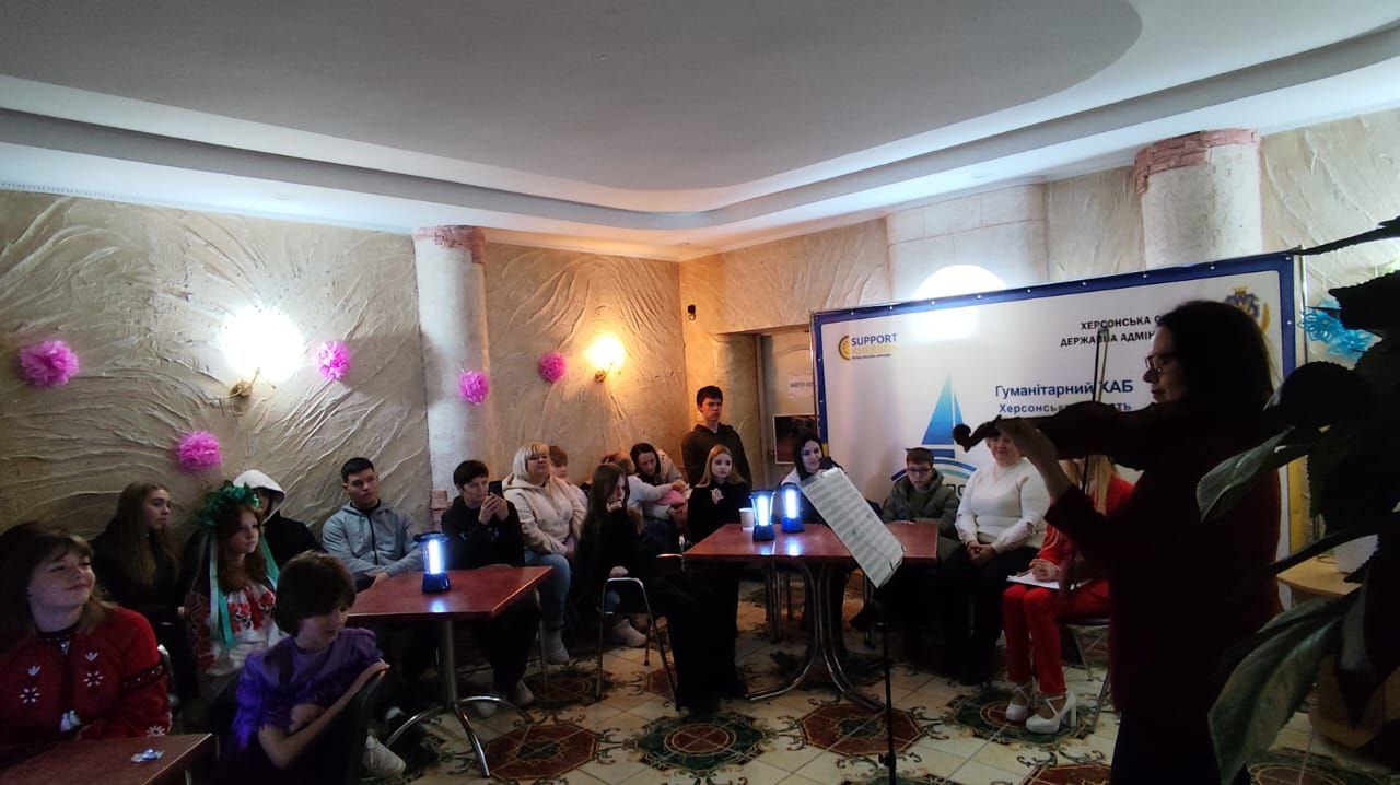 Сцена, де жінка грає на скрипці перед аудиторією, що складається переважно з молоді, яка сидить за столами. Подія відбувається у приміщенні Гуманітарного хабу Херсонської області, про що свідчить банер на задньому плані.