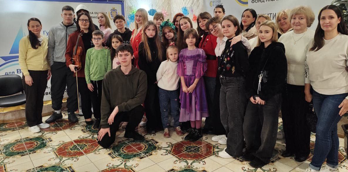 Групове фото, на якому близько двадцяти дітей, підлітків та дорослих стоять або сидять на колінах у приміщенні волонтерського хабу, посміхаючись. На задньому плані видно рекламний банер.