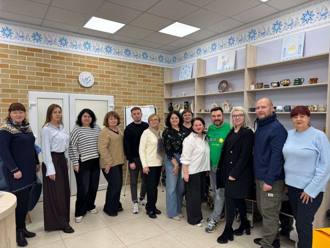 Групова фотографія одинадцяти дорослих людей (чоловіків та жінок), які стоять у приміщенні зі світлими цегляними стінами та полицями з декоративними виробами.