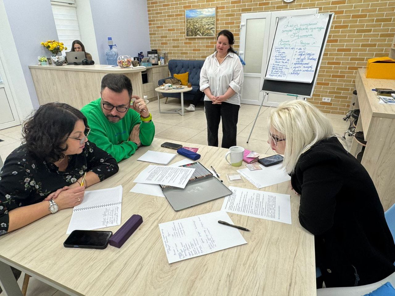 Троє людей сидять за столом, працюючи з документами та блокнотами, поки четверта жінка стоїть позаду них, спостерігаючи за роботою групи в приміщенні для тренінгів.
