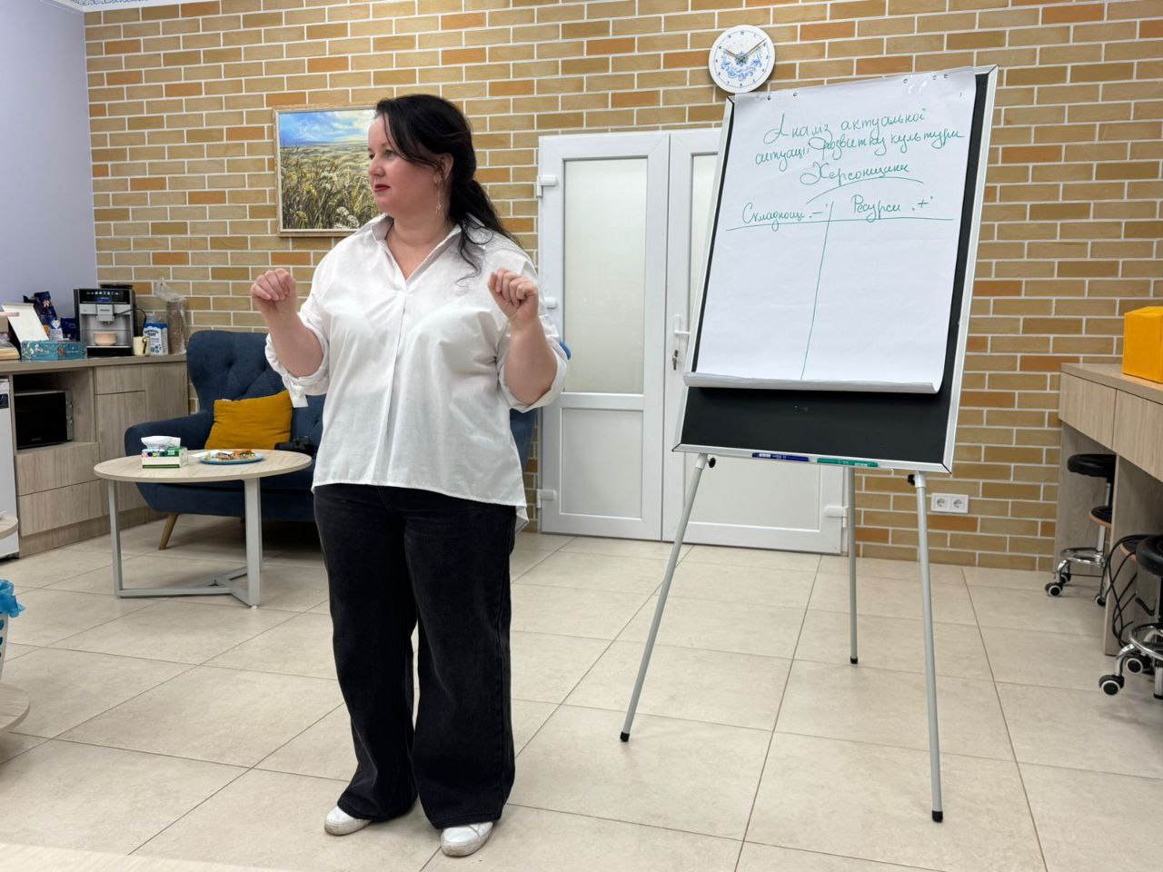 Жінка в білій сорочці та чорних штанах стоїть перед фліпчартом, на якому написано текст. Вона жестикулює руками, ніби пояснює матеріал під час презентації чи тренінгу.