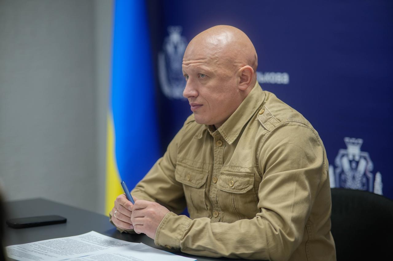 Портрет лисого чоловіка в бежевій військовій сорочці, який сидить за столом, тримаючи ручку. На задньому плані синьо-жовтий прапор України та синій банер з логотипом адміністрації.