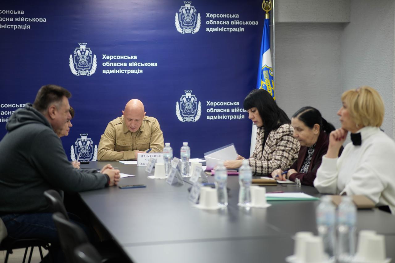 Група людей, включно з чоловіком у військовій формі та жінками, сидять за довгим столом під час наради. На синьому фоні позаду повторюється логотип Херсонська обласна військова адміністрація.