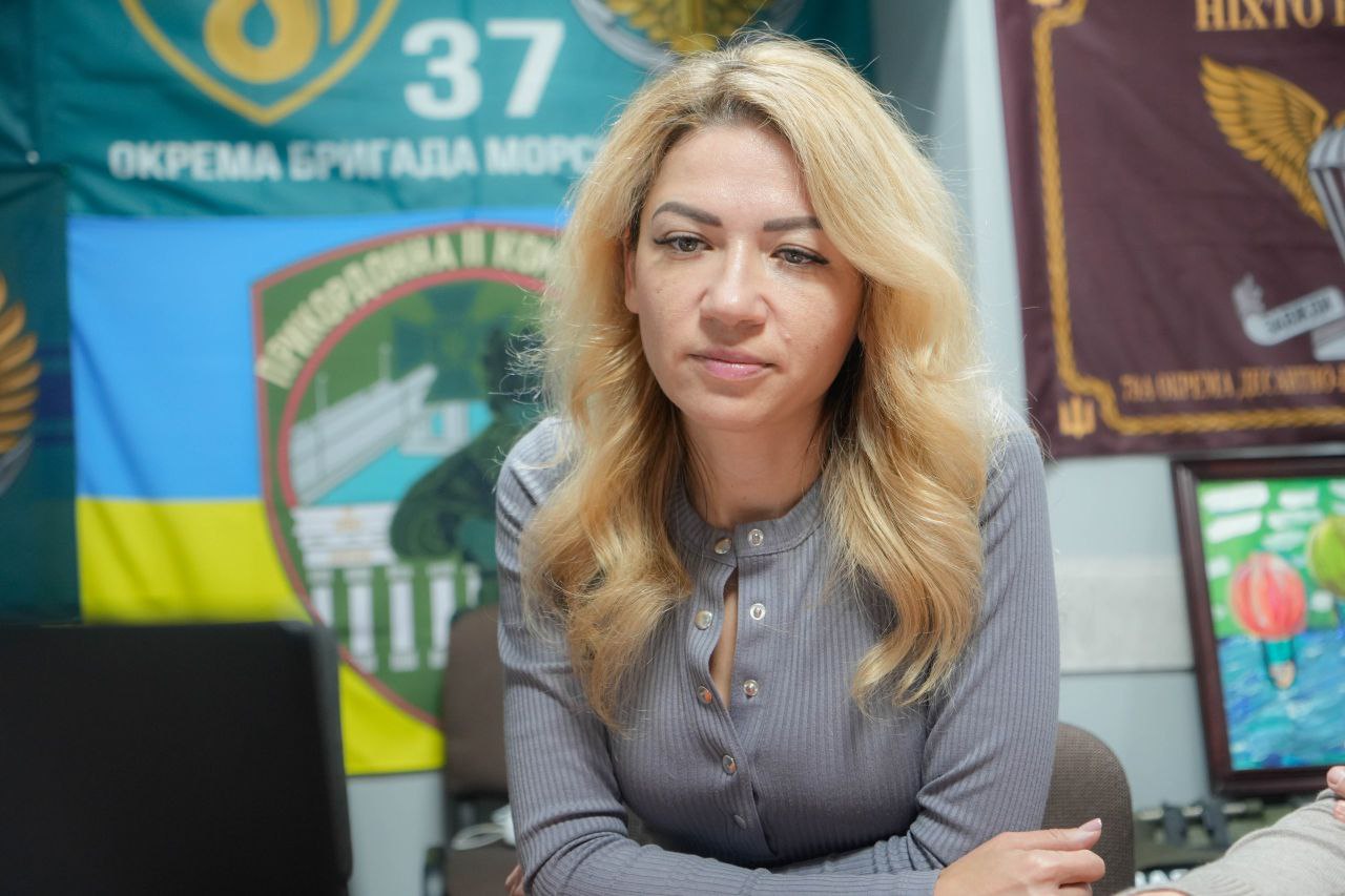 Жінка з довгим світлим волоссям сидить за столом у сірому светрі з ґудзиками. На задньому плані видно військові прапори та український прапор.