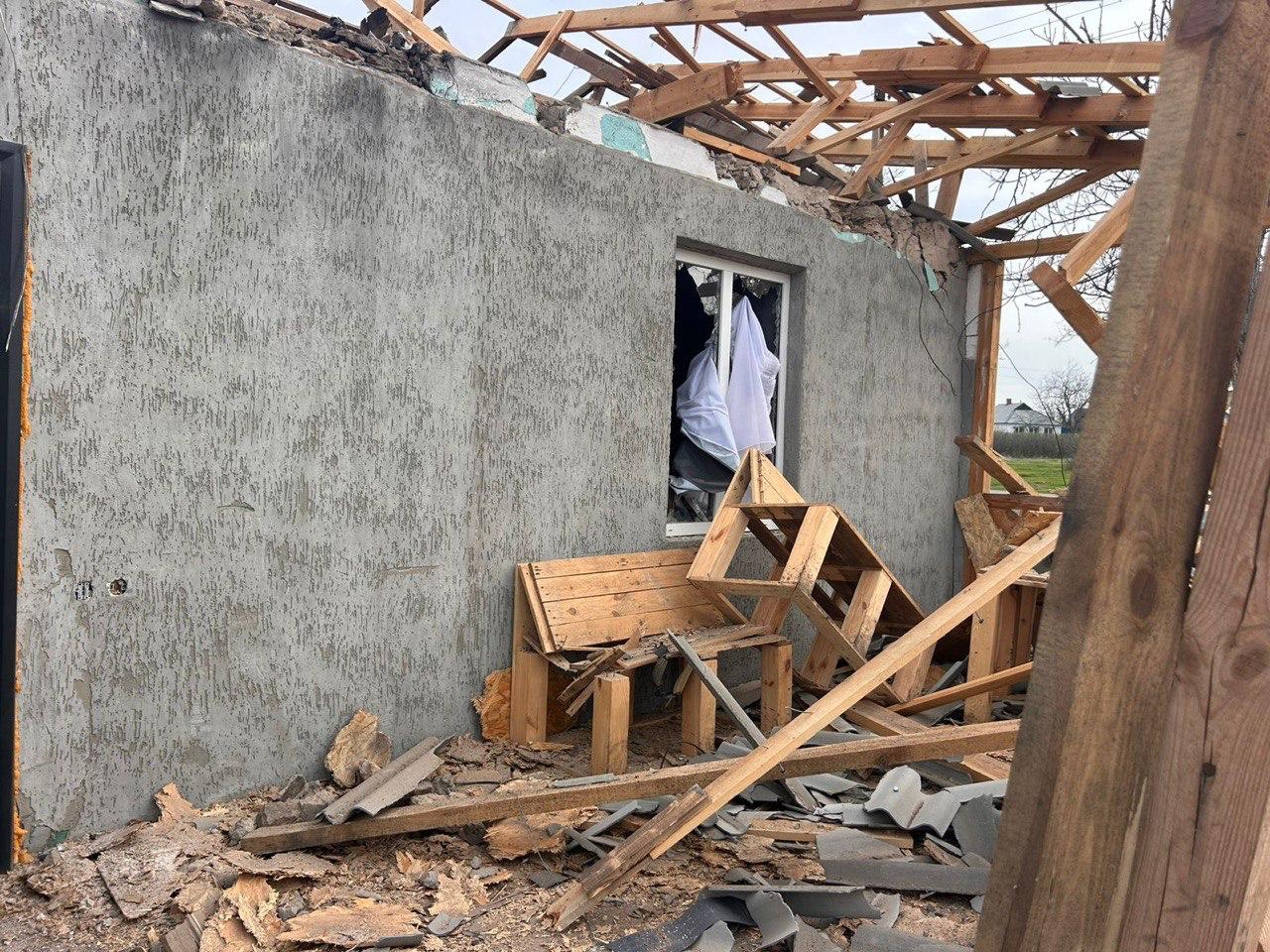 Крупний план частини зруйнованого будинку: сіра стіна з пошкодженим вікном, з якого висить біла тканина, відсутній дах (видно дерев'яні крокви). Навколо будинку лежать уламки шиферу, дошки та зламані дерев'яні конструкції, зокрема лавка.