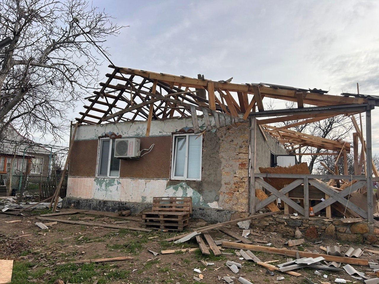 Фасад сильно пошкодженого будинку. Будинок без частини даху, видно лише дерев'яні крокви. Частина фасаду облущена, видно два вікна та кондиціонер. Навколо розкидані будівельні уламки та дошки.