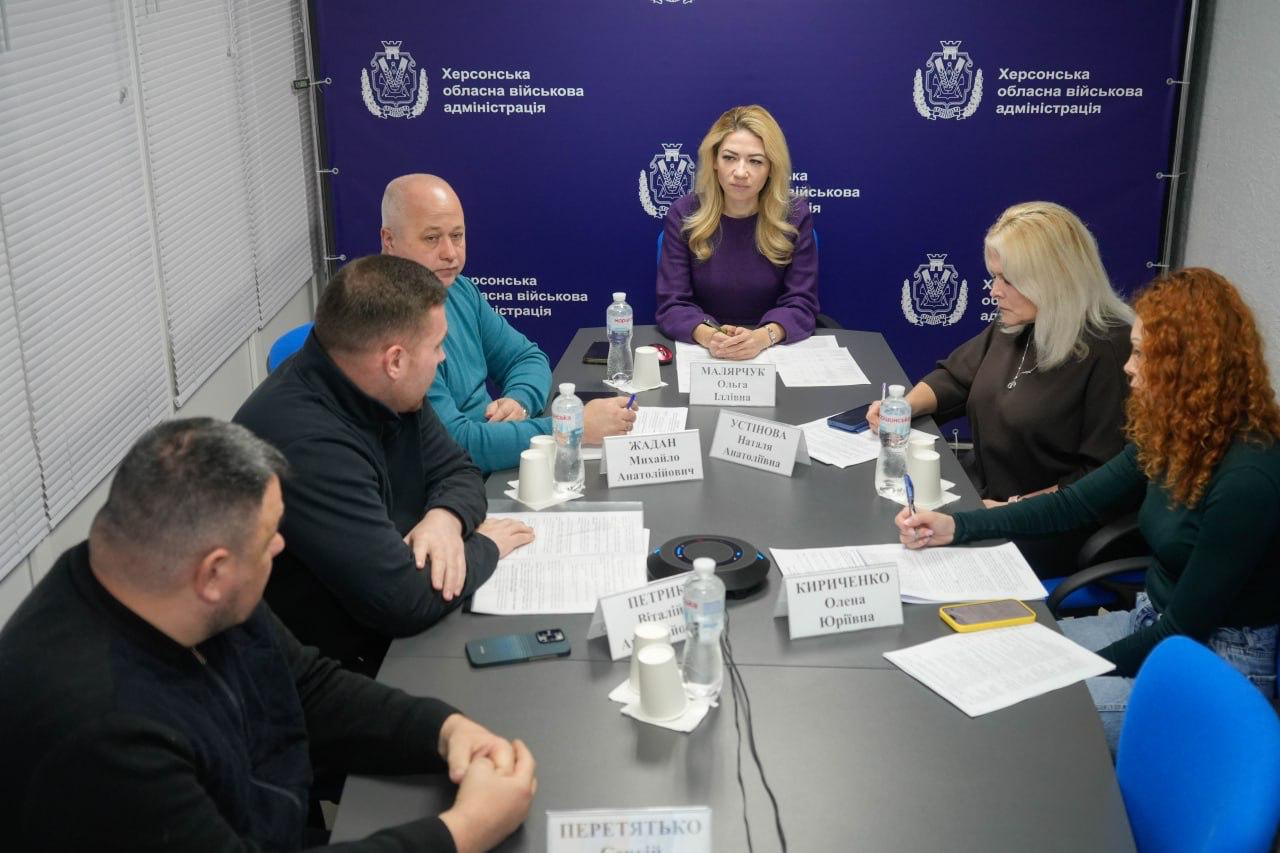 Нарада посадових осіб Херсонської обласної військової адміністрації (ХОВА) за великим столом. Учасники обговорюють документи, перед ними стоять таблички з прізвищами.