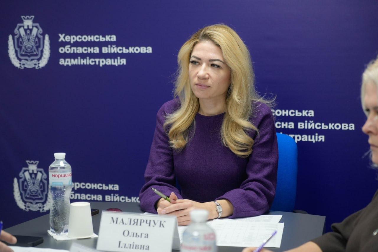 Жінка зі світлим волоссям сидить на нараді, тримаючи ручку. На задньому плані видно логотипи Херсонської обласної військової адміністрації.