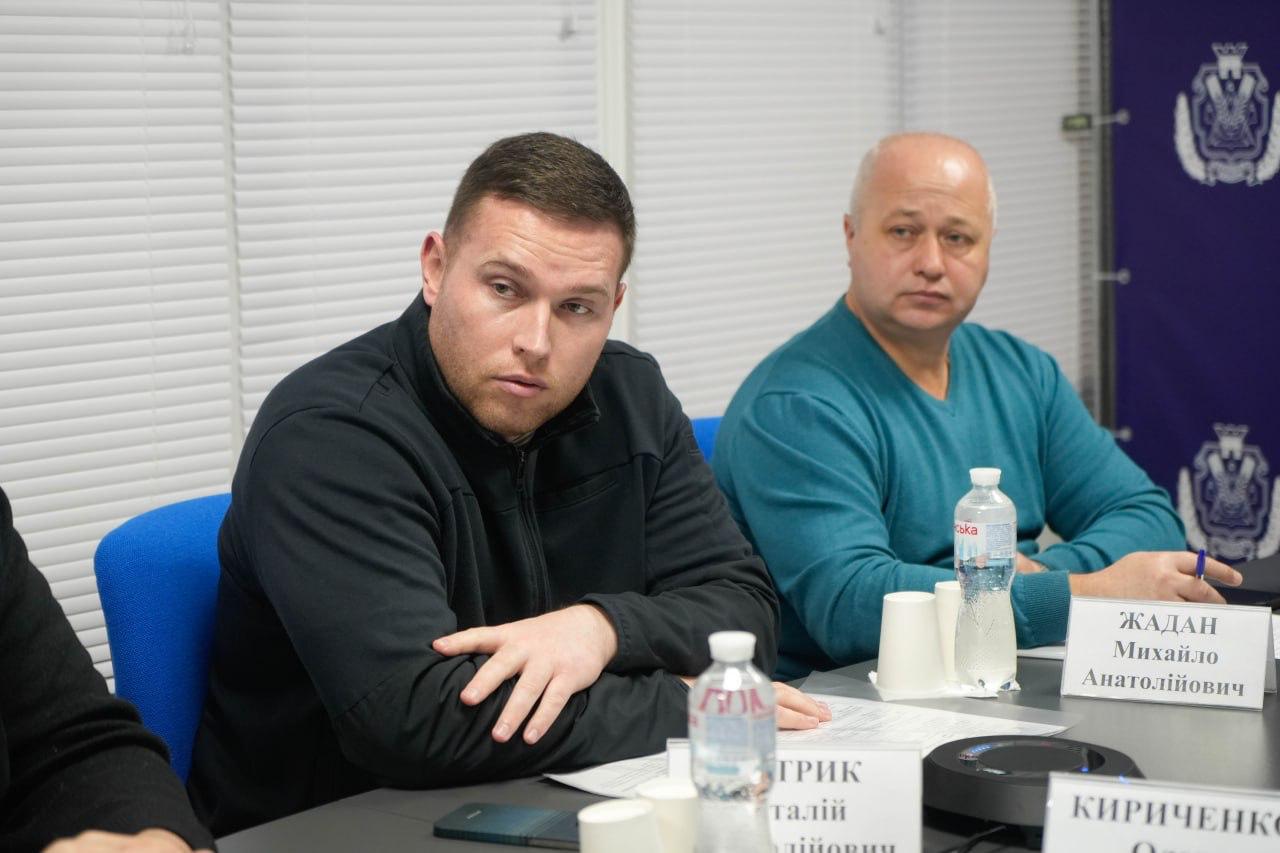 Двоє чоловіків на нараді в Херсонській ОВА. На передньому плані — чоловік у темній кофті, який сидить, спершись на стіл. Поруч із ним — лисий чоловік у бірюзовому светрі.